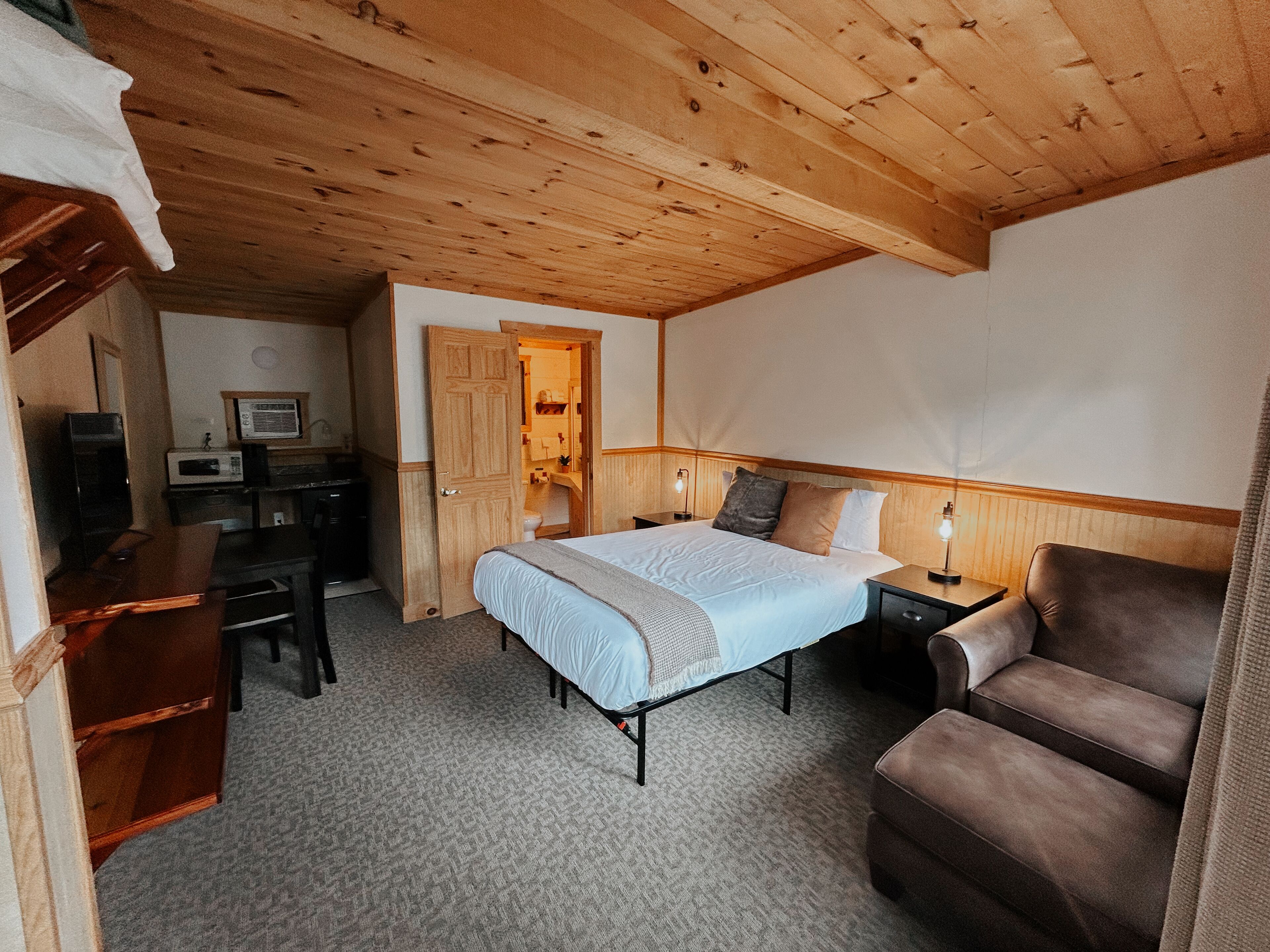 Un dormitorio con cama, escritorio, silla y baño visible a través de una puerta abierta.