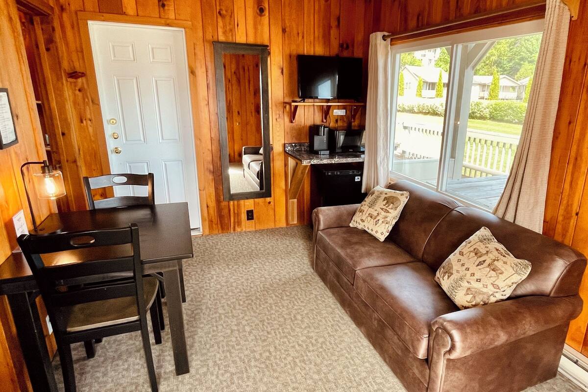 A cozy room with a brown leather sofa, a dining table with chairs, a kitchen area with a TV, and a large window offering a view of the outside.