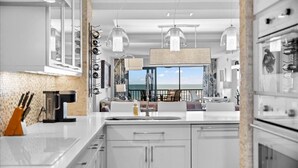 Une cuisine moderne avec des meubles blancs, un grand îlot central, des luminaires suspendus et une vue sur l’océan grâce à une grande fenêtre.
