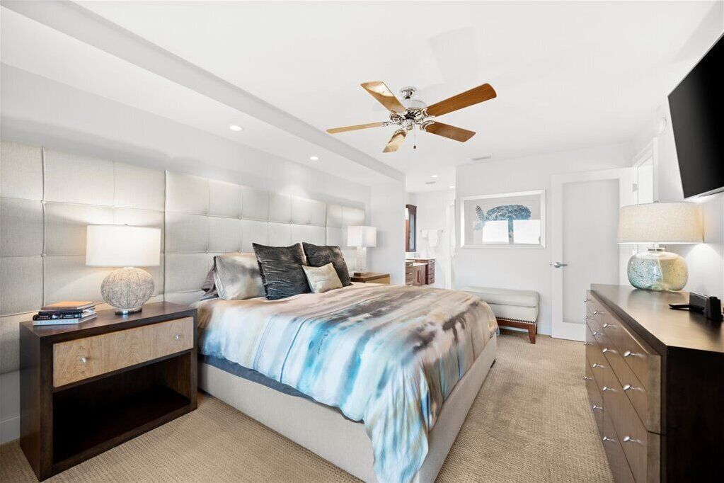 A modern bedroom with a large bed, a ceiling fan, a bedside table with a lamp, and a dresser.