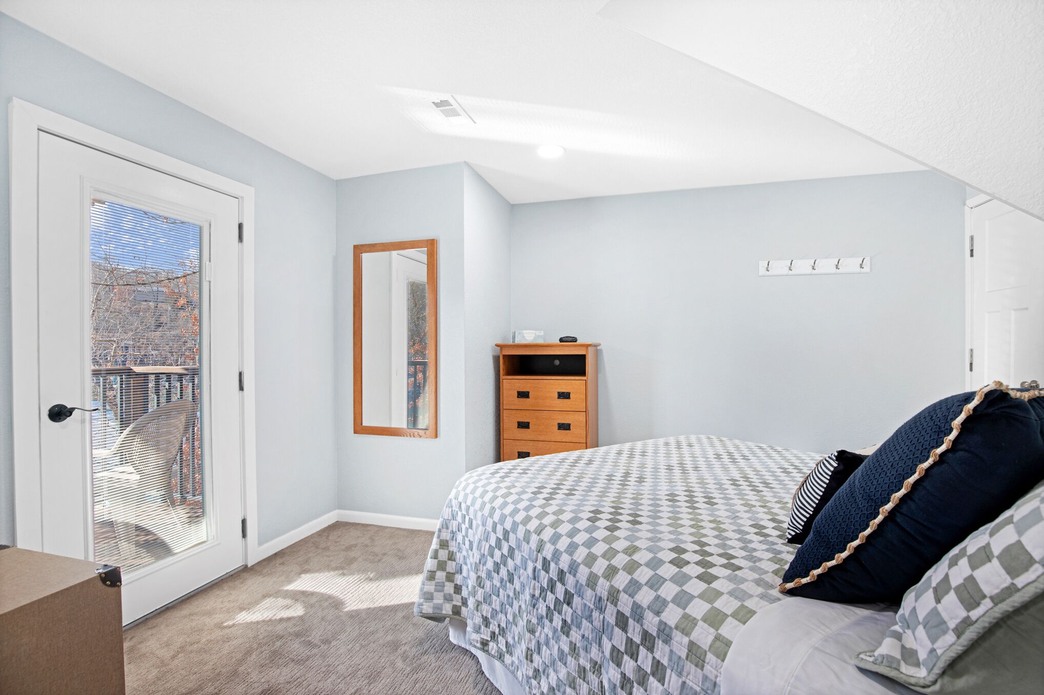 A bedroom with a checkered bedspread, a wooden nightstand, a mirror, and a door leading to a balcony.
