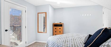 A bedroom with a checkered bedspread, a wooden nightstand, a mirror, and a door leading to a balcony.