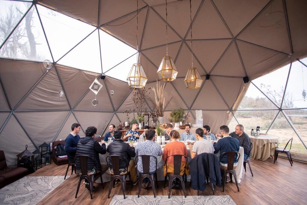A group of people dining inside a geodesic dome tent with hanging pendant lights and a wooden floor.