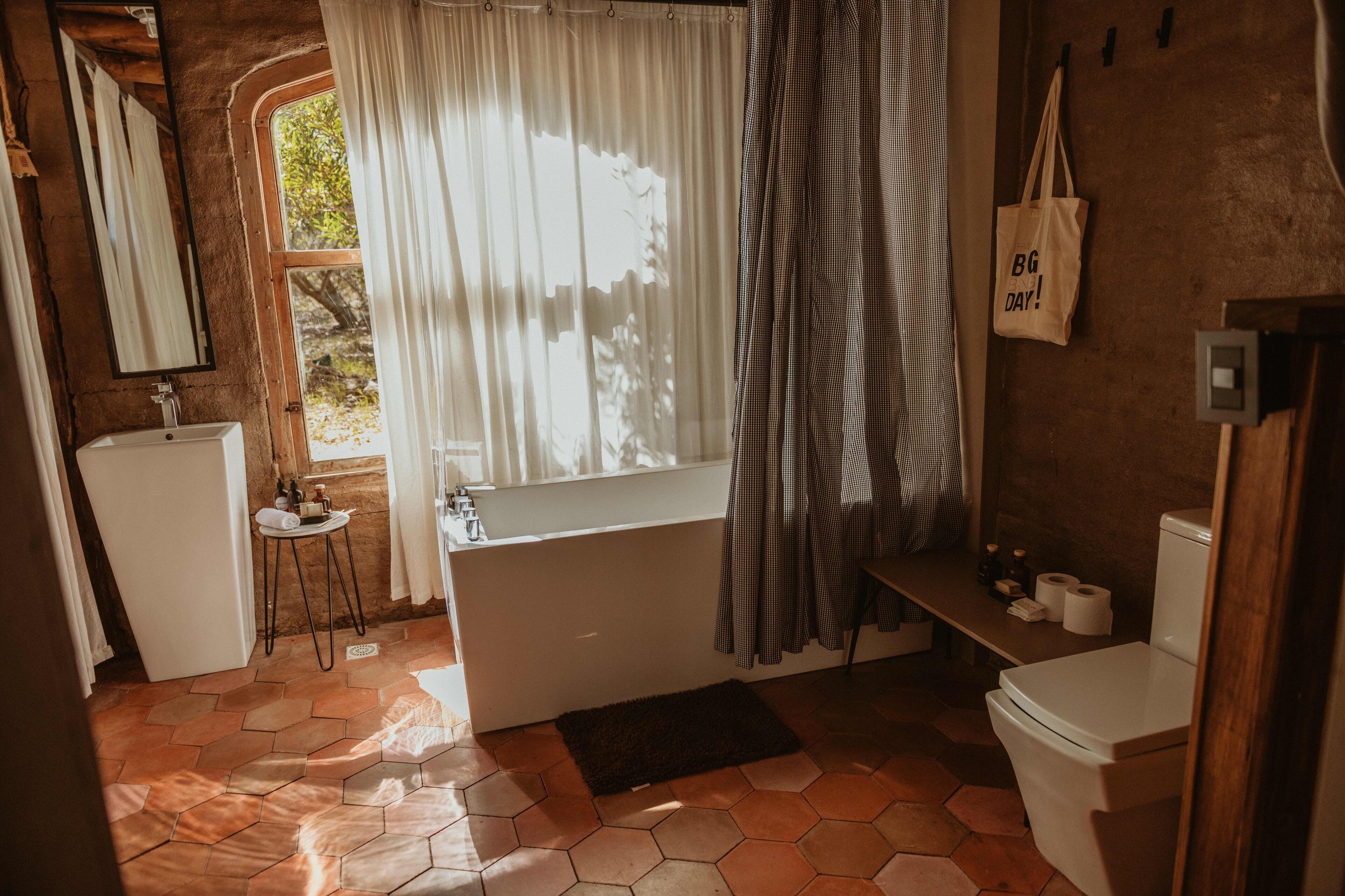 Ein Badezimmer mit Badewanne, Waschbecken und Toilette. Es gibt ein Fenster mit Vorhängen, einen Spiegel und einen Handtuchhalter.