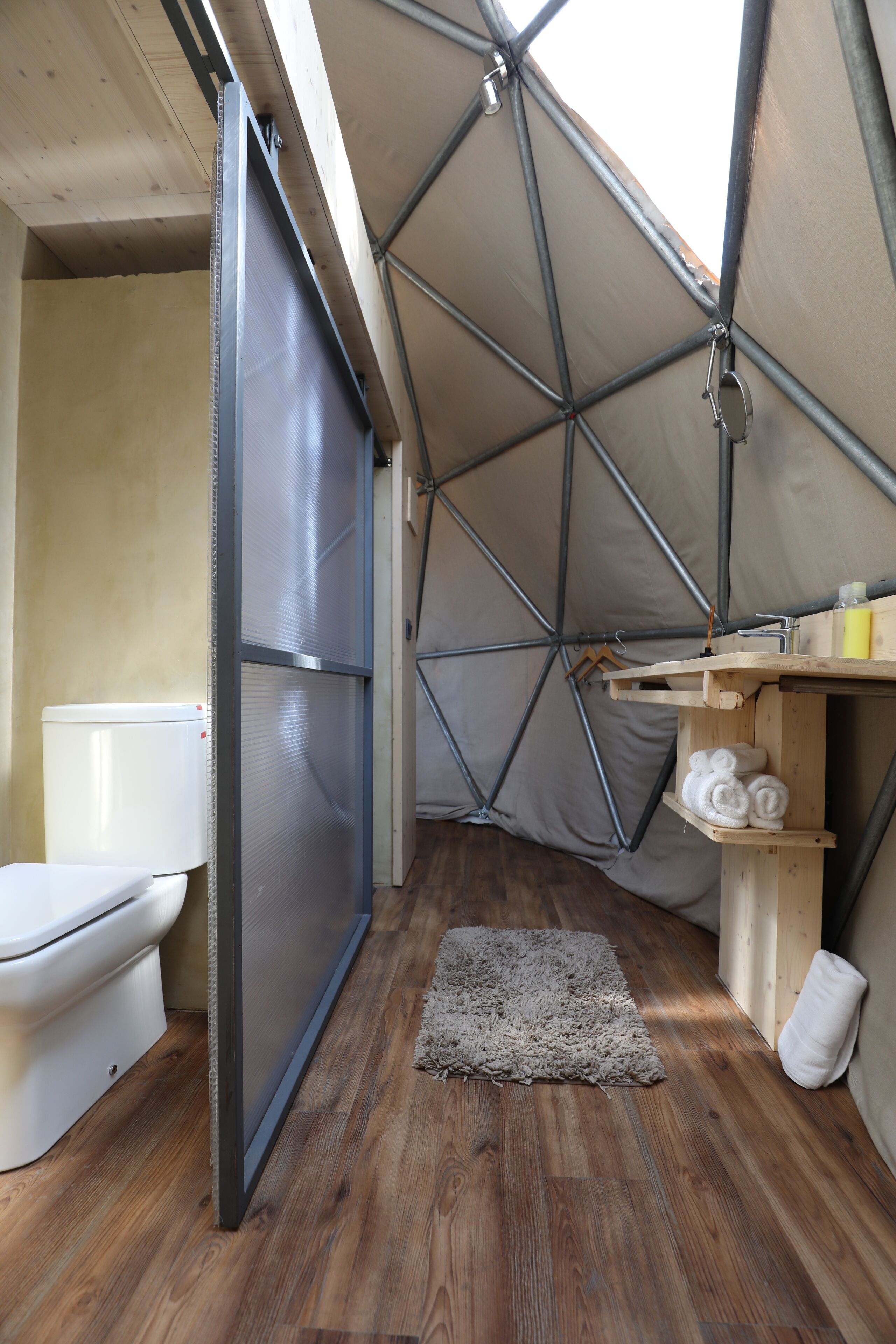 A compact, modern bathroom with a wooden floor, a toilet, a shower area, and a small shelf with towels.