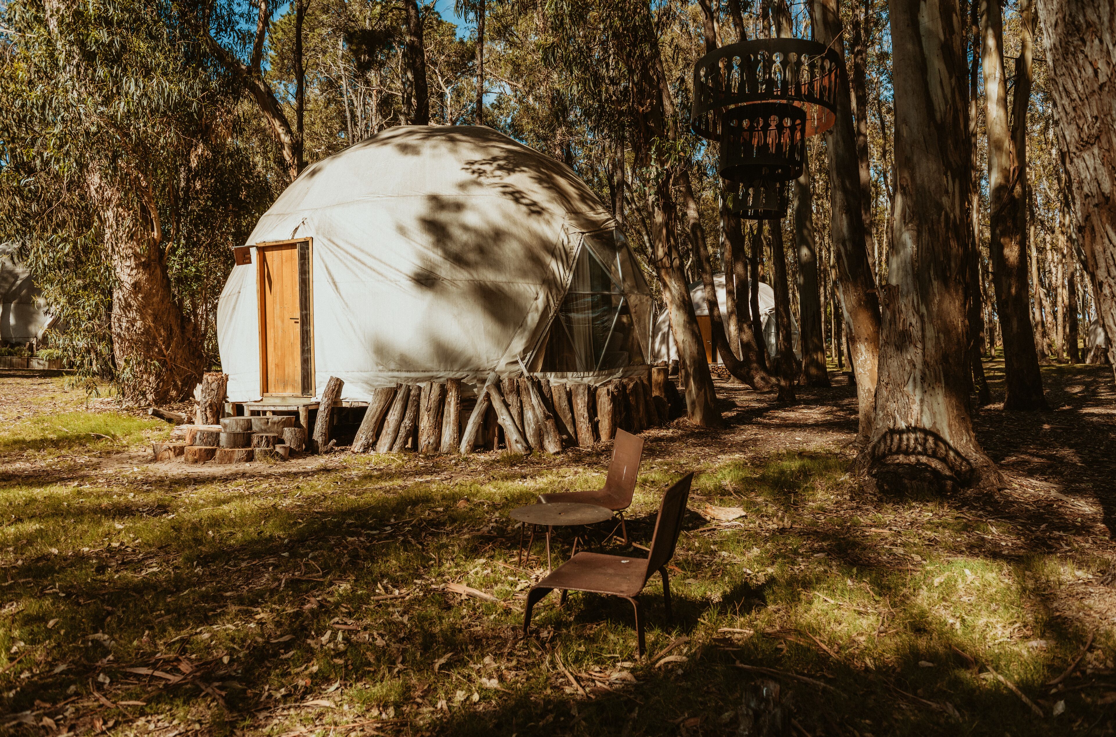 A rustic yurt with a wooden door, surrounded by trees and a small wooden table with chairs.