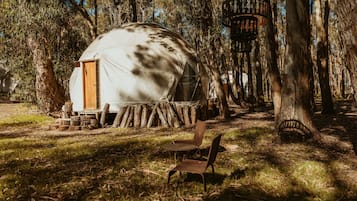 Una yurta rústica con puerta de madera, rodeada de árboles y una pequeña mesa de madera con sillas.