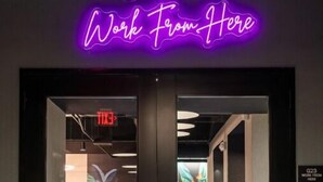 Ein Hotel Eingang mit einem Neonlichtschild, das "Work From Here" zeigt, und Blick in einen Lobybereich mit Sitzgelegenheiten und einem Wandgemälde.
