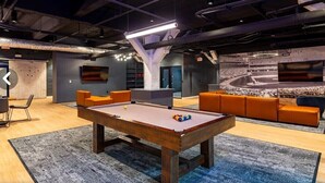 Ein Billardtisch in einem modernen Lounge-Bereich mit orangefarbenen Sitzgelegenheiten und einem großen Bildschirm, der ein Stadion zeigt.