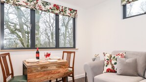 A dining area with a wooden table and chairs, a sofa, and a window with a floral curtain. - Herdwick Cabin (Penrith)
