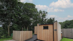 A wooden cabin with a small window and a door, surrounded by a gravel path and a wooden fence. - Herdwick Cabin (Penrith)