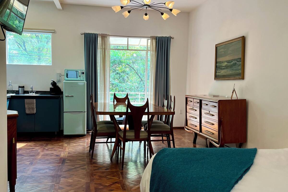A compact room with a dining table, chairs, a bed, a kitchenette, and a large window with curtains.
