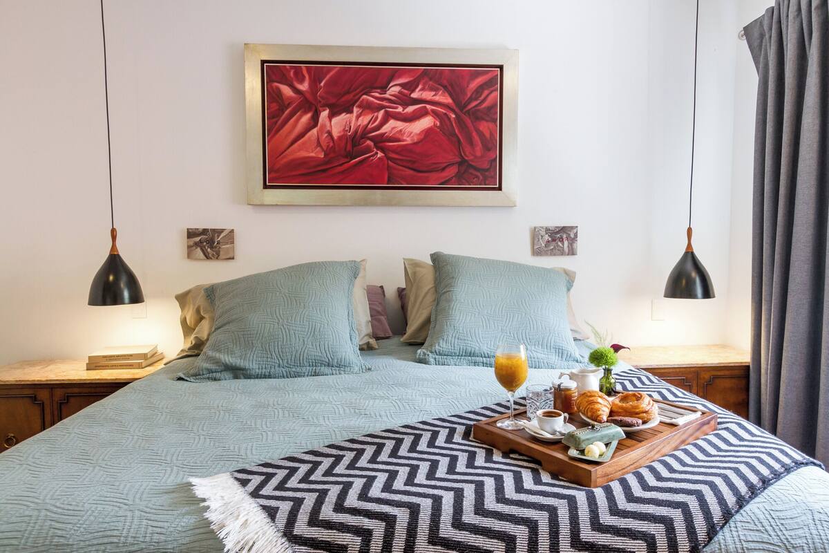 A neatly arranged bedroom with a bed, pillows, a tray of breakfast items, and artwork on the wall.