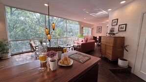 Un amplio salón con una mesa de centro de madera, un sofá y una mesa de comedor. Grandes ventanales ofrecen vistas al exterior.