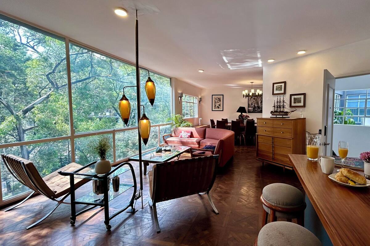 A spacious living area with a large window, a dining table with chairs, a sofa, and a wooden cabinet.
