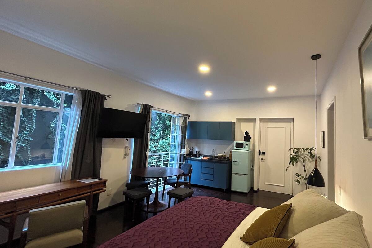 A compact room with a bed, a desk, a chair, a small kitchenette, and a window with curtains.