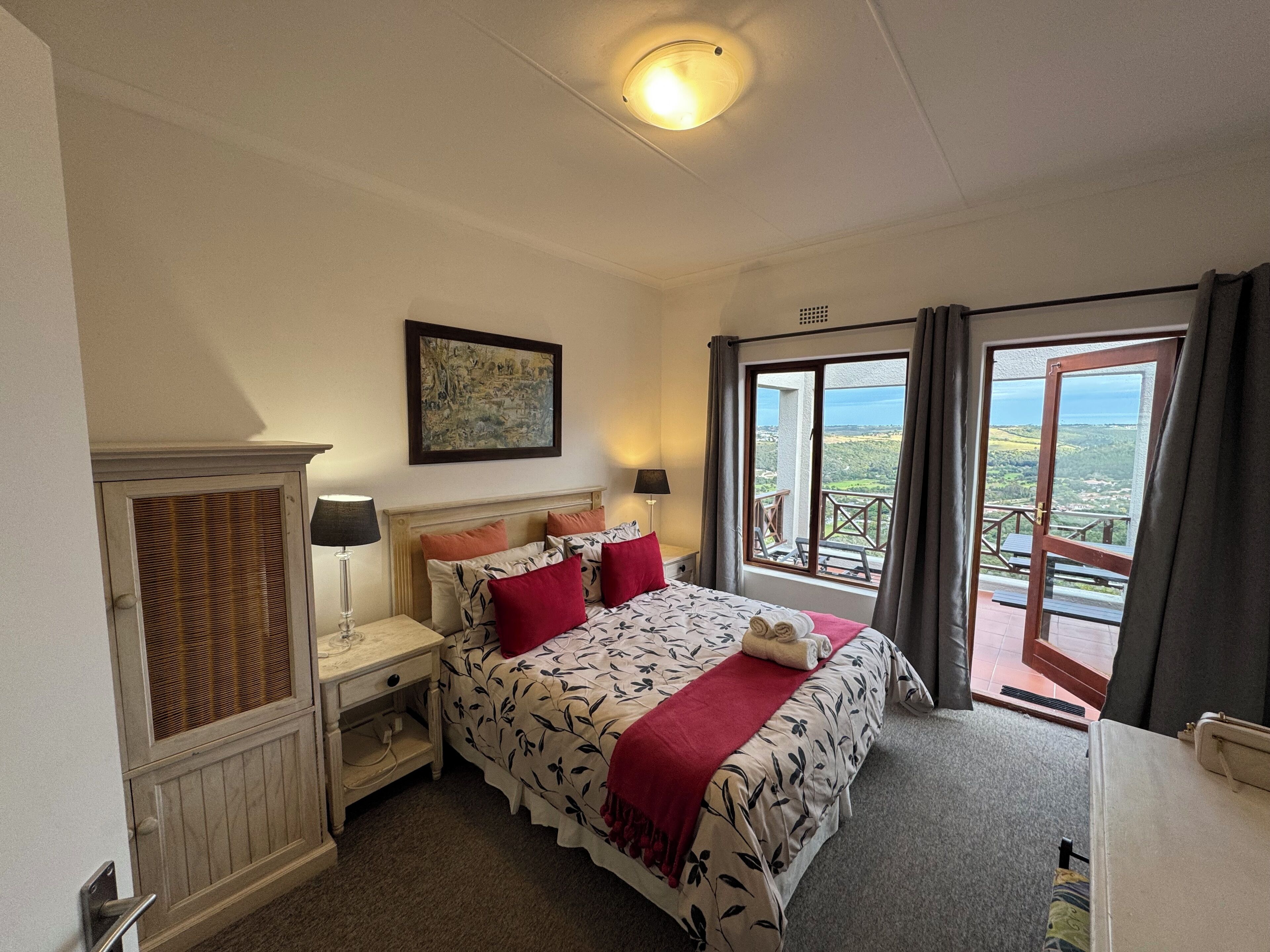 A bedroom with a bed, bedside tables, a nightstand, a painting, and a balcony with a view.