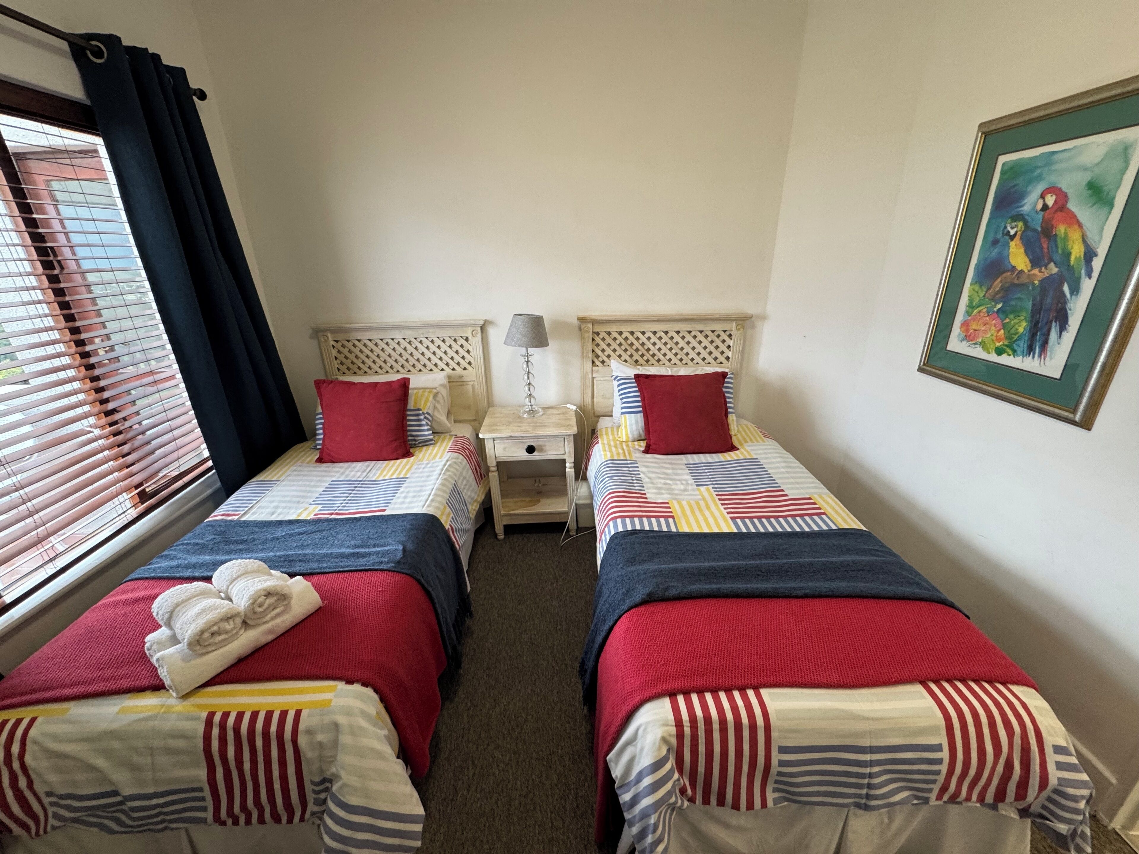 A hotel room with two single beds, each with a red pillow and a striped blanket. There is a nightstand with a lamp between the beds, and a painting on the wall.