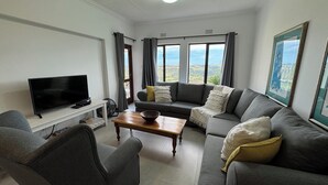 A living room with a TV, sofa, and large windows offering a view of the landscape. - Castleton 19B Family friendly  apartment, in a secure resort, in Plettenberg Bay (Plettenberg Bay)