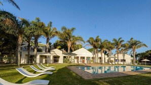 A poolside area with lounge chairs, a swimming pool, and palm trees. - Castleton 19B Family friendly  apartment, in a secure resort, in Plettenberg Bay (Plettenberg Bay)