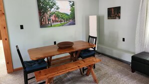 A dining area with a wooden table, two blue cushioned chairs, a bench, and a painting of a street scene on the wall.
