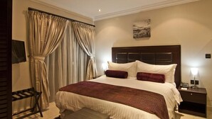 A hotel room with a large bed, a headboard, a bedside table with a lamp, and a window with curtains. - Alheri Royal Apartment (Abuja)