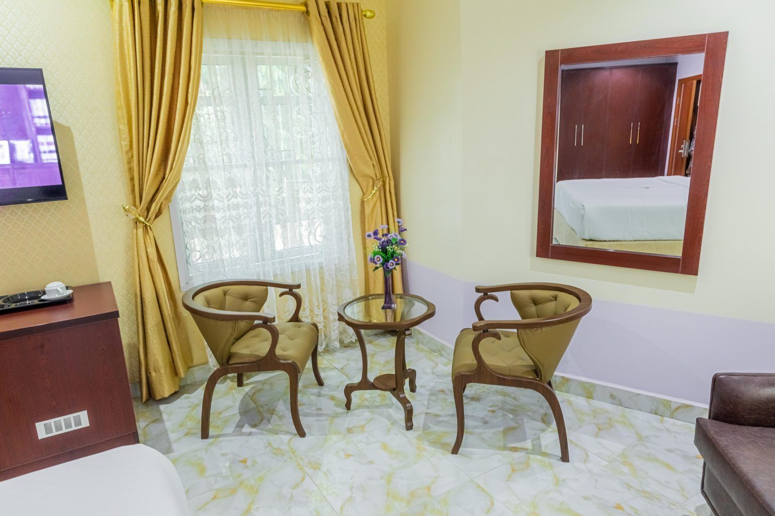 A hotel room with a bed, a desk with a TV, two armchairs, a small round table with a vase, and a mirror on the wall.