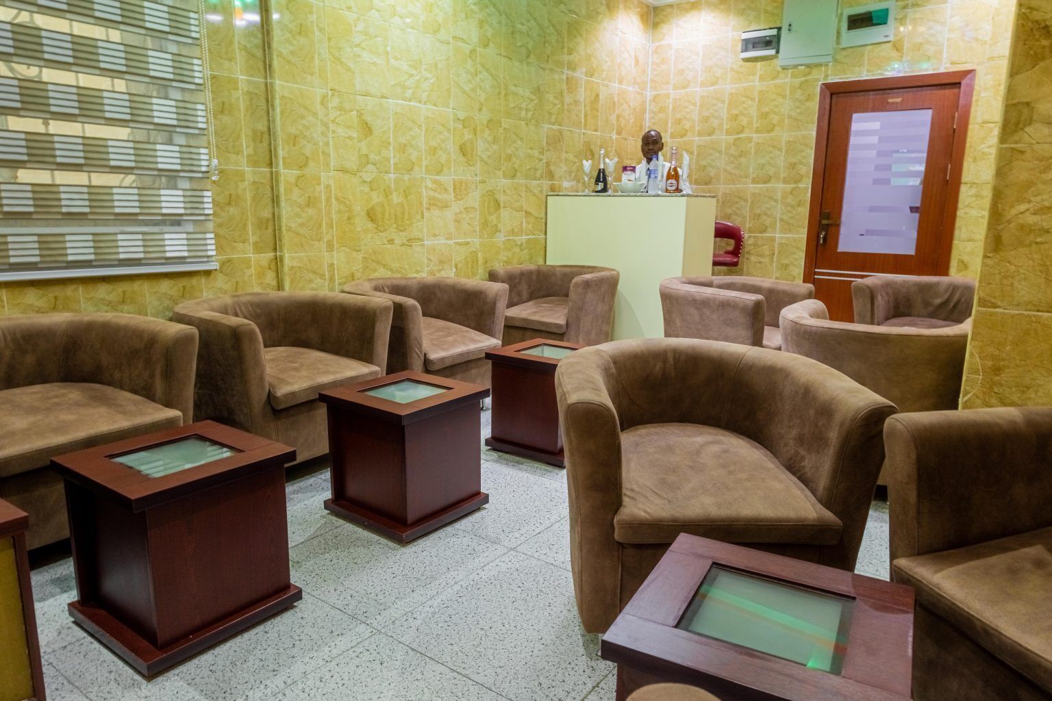 A room with multiple brown armchairs and small wooden tables, a reception desk with a counter, and a tiled wall.