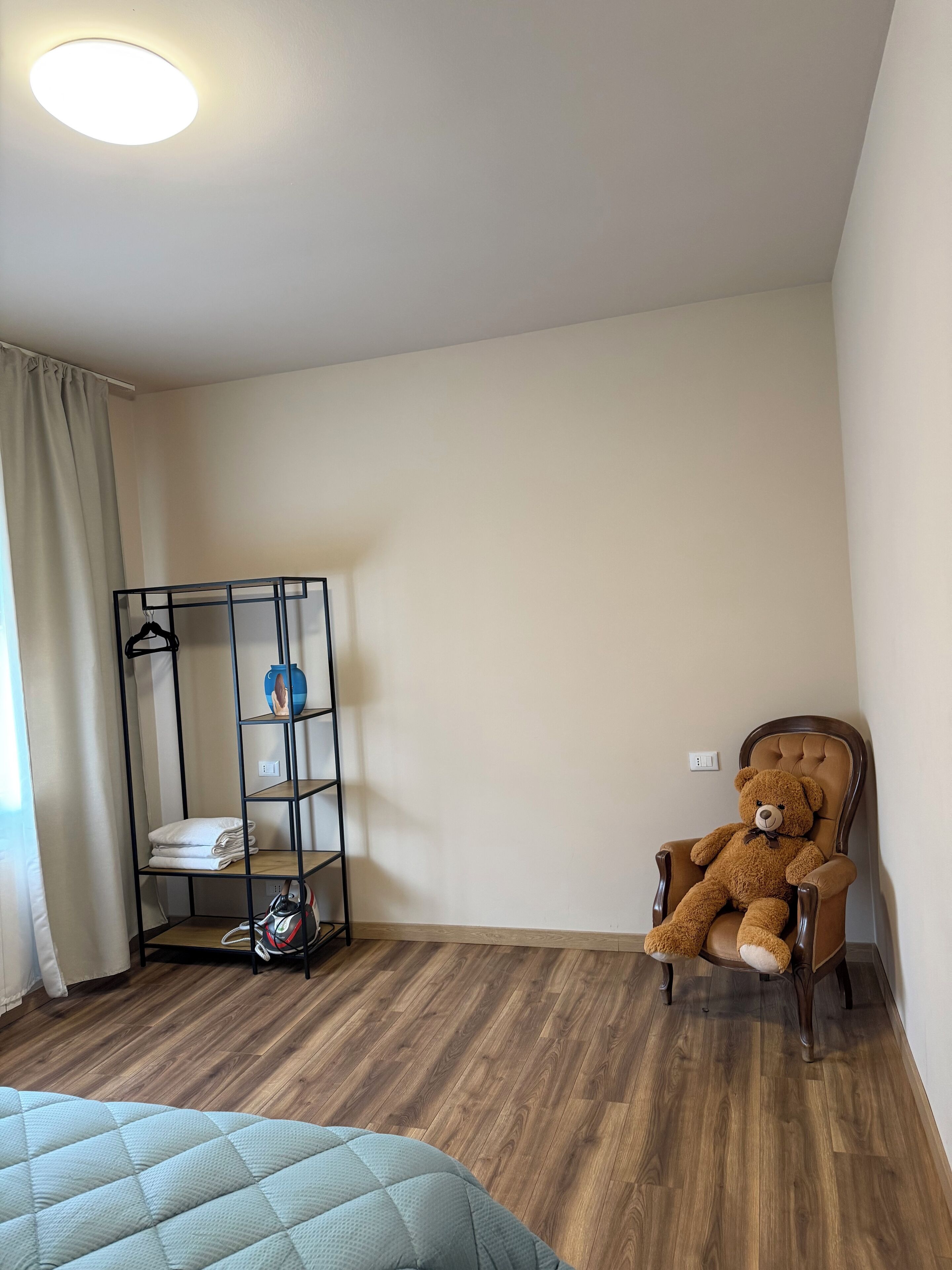 A room with a wooden floor, a bed with a quilt, a chair with a teddy bear, a metal shelf unit with folded towels and a vase, and a ceiling light.