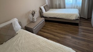 A modern hotel room with two beds, a wooden floor, and a large window with sheer curtains.