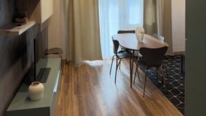 A modern, well-lit room with a wooden floor, a dining table with chairs, a small shelf with a vase, and a window with curtains.