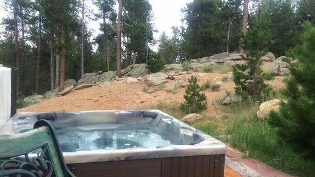 A hot tub with a wooden deck and metal bench, surrounded by a natural landscape with trees and rocks. - Mtn Retreat: Hike ,Hot Tub, Explore CO, Ski, Relax (Golden)