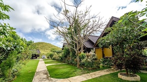 A pathway leading to a house with a yellow exterior, surrounded by lush greenery and a hill in the background. - Deluxe Bungalow with Bathtub, Pini Sentana Village (Nusa Penida)