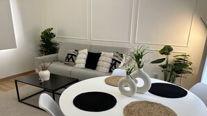 En moderne stue med et rundt hvidt bord, to hvide vaser med planter, en grå sofa med mønstrede puder og et lille sort sofabord.