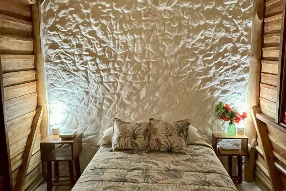 Um quarto aconchegante com teto de madeira, uma cama com roupa de cama floral, duas mesas de cabeceira e um criado-mudo com um vaso de flores.