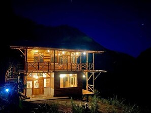 A two-story wooden cabin with a balcony, illuminated windows, and a staircase leading to the entrance. - Eco-Finca Hostal Santorum (Vilcabamba)
