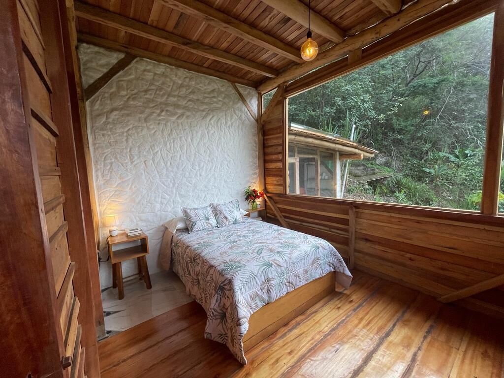 Una camera da letto accogliente con soffitto in legno e un'ampia finestra con vista su una zona boschiva.