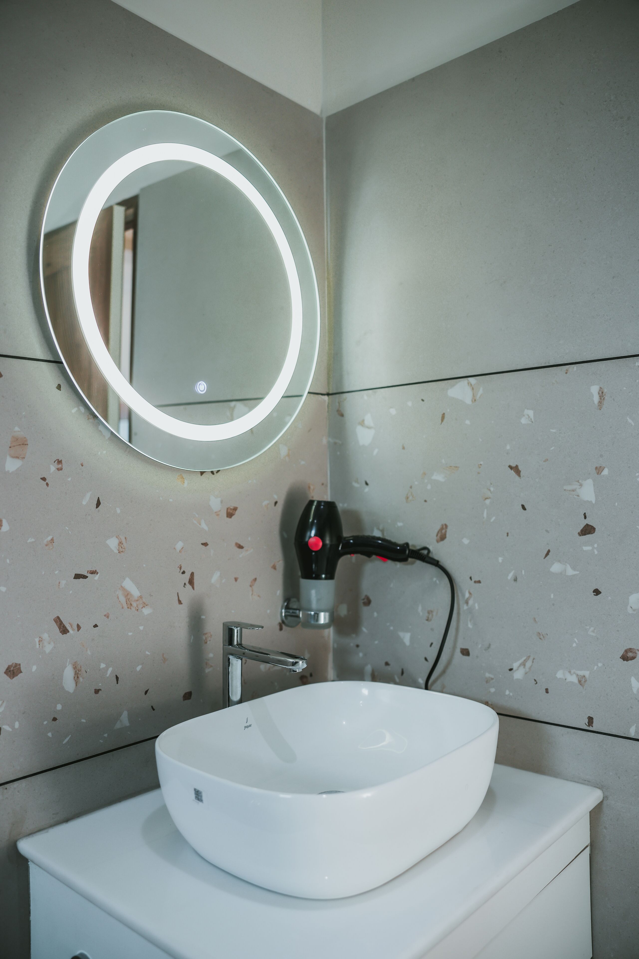 A bathroom with a white sink, a round mirror with a light, and a hairdryer on the wall.
