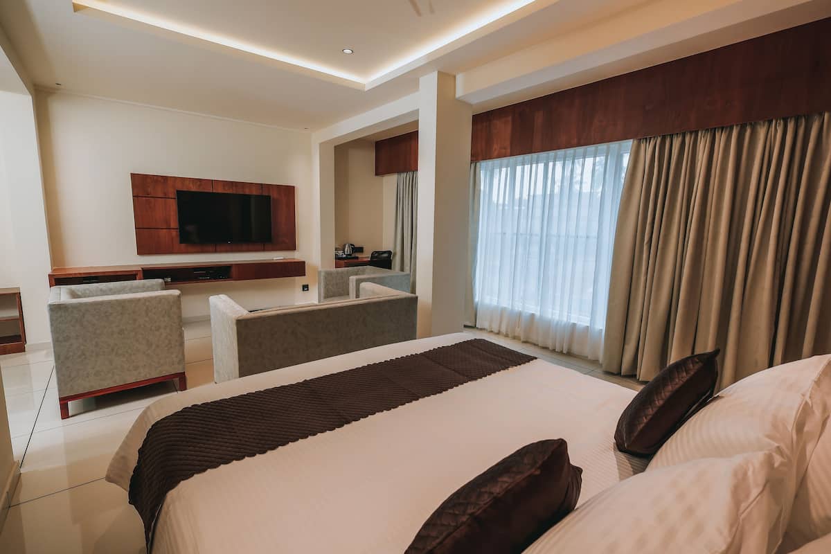 Una habitación de hotel moderna con una cama grande, un televisor de pantalla plana en la pared, una zona de estar con sofá y sillones, y un ventanal con cortinas.