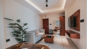 Un salon moderne avec un canapé, une table basse et une grande plante. Un téléviseur est fixé au mur et un ventilateur de plafond est installé.