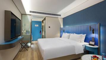 A modern hotel room with a large bed, a TV, a bedside table with a lamp, and a small round table with fruit.