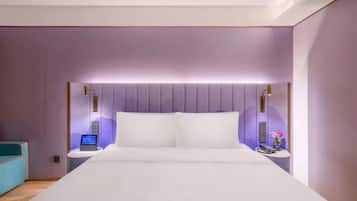 Una habitación de hotel moderna con una cama grande, una mesita de noche con un teléfono y un jarrón de flores, y un panel de control en la pared.