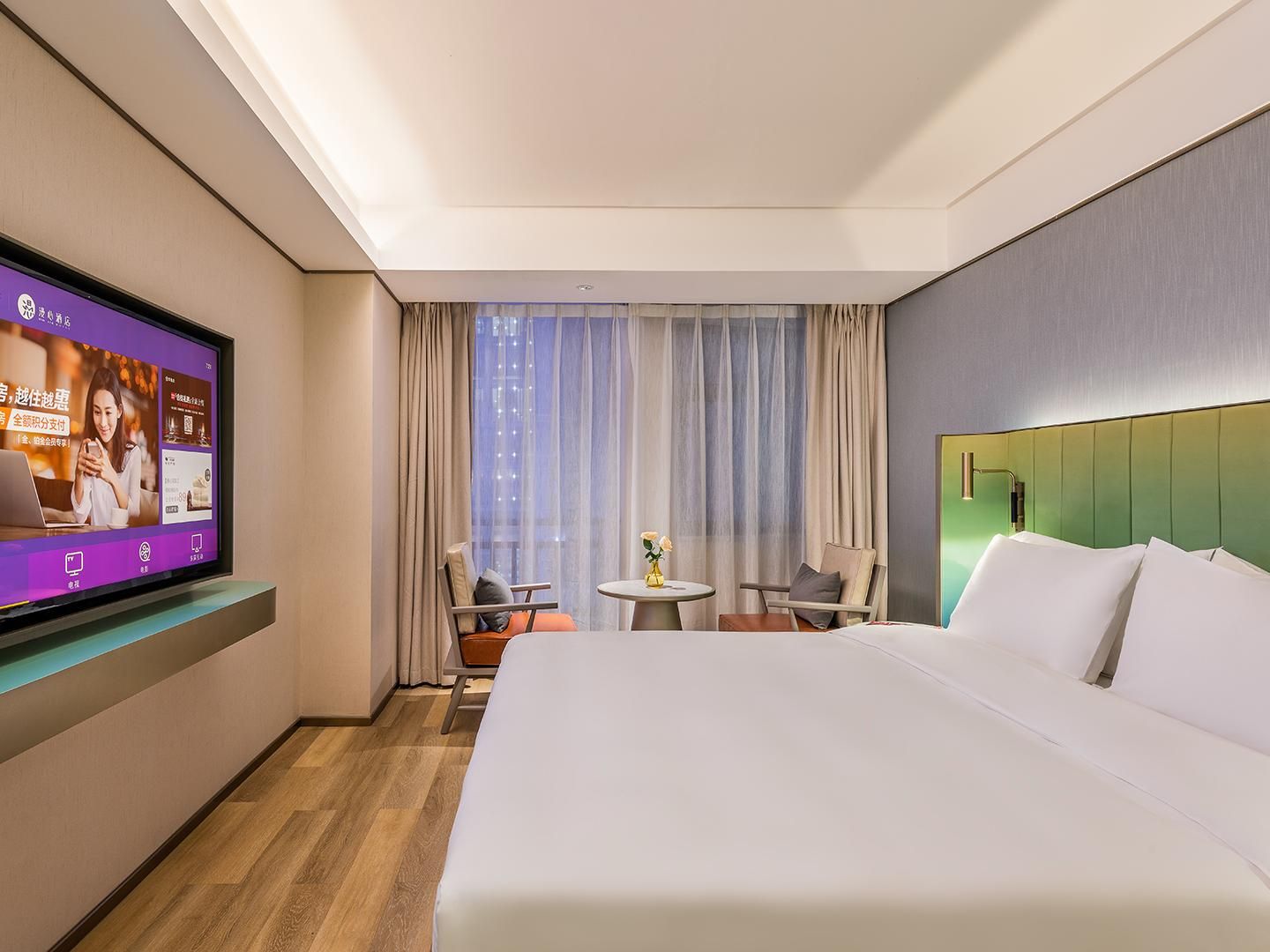 A hotel room with a bed, a TV mounted on the wall, a small table with chairs, and a window with curtains.