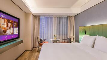 A hotel room with a bed, a TV mounted on the wall, a small table with chairs, and a window with curtains.