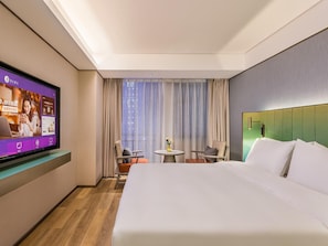 Habitación de hotel con cama, televisor montado en la pared, una mesita con sillas y una ventana con cortinas.