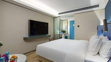 A hotel room with a bed, a TV mounted on the wall, a small round table with fruit and drinks, and a bathroom visible through a glass door.