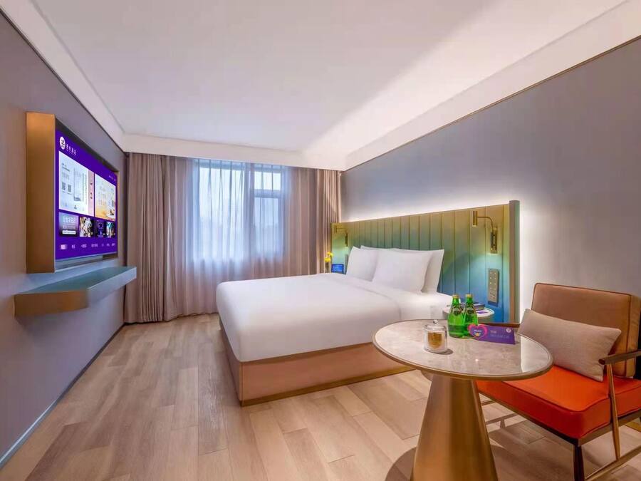 A hotel room with a bed, a round table with a chair, a TV screen, and a window with curtains.