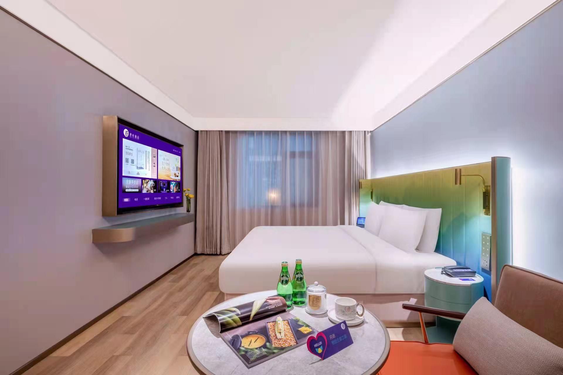 A hotel room with a bed, a TV mounted on the wall, a bedside table with a lamp, a small round table with a magazine, and a bottle of water.