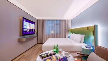 A hotel room with a bed, a TV mounted on the wall, a bedside table with a lamp, a small round table with a magazine, and a bottle of water.