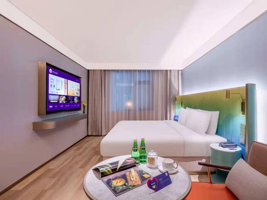 A hotel room with a bed, a TV mounted on the wall, a bedside table with a lamp, a small round table with a magazine, and a bottle of water.